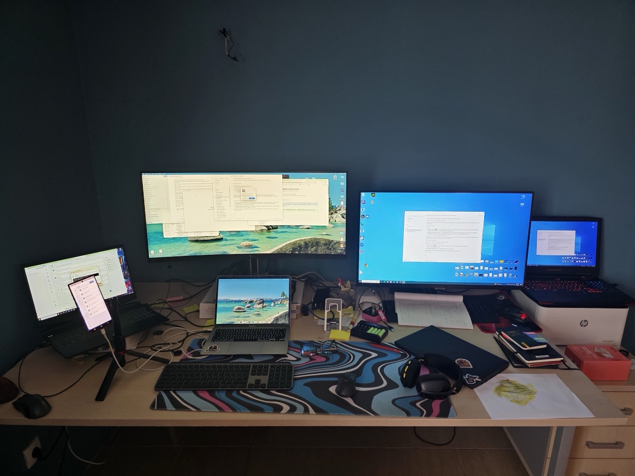 Wide bench shot of four benchmark machines on a home-office desk, with a child's drawing of a flower in the bottom-right corner.