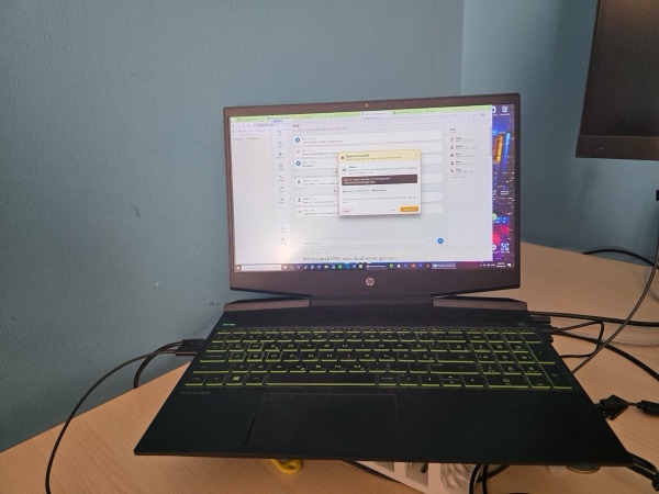 An HP gaming laptop, lid open, green keyboard backlight, running a chat application on a cluttered home-office desk.
